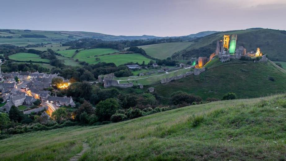 A long exposure shot of Corfe Castle lit up green and yellow with the village lights below