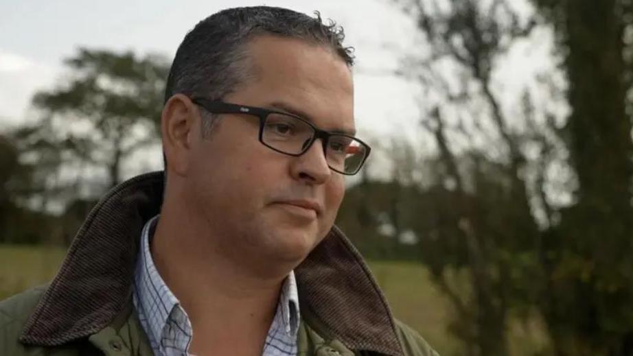 Marcus Calvani photographed from the shoulders up. Wearing a green coat, check shirt and glasses. He has short black hair and black framed glasses. A countryside scene is visible behind him. 