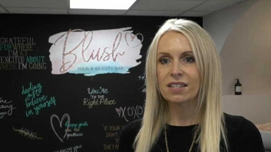 A woman with blonde hair is wearing a black top and a gold necklace, she is stood in front of a black wall which has the logo of her company on it and a number of affirmations in the style of a chalk board.