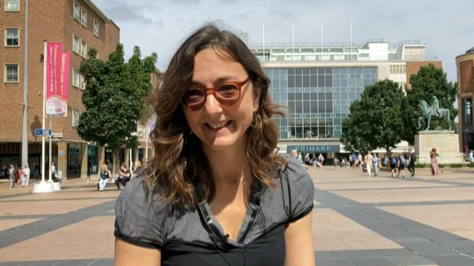 A woman is smiling at the camera, she is wearing glasses and has shoulder-length wavy brown hair. She is wearing a gret and black shirt and is sitting in Coventry city centre