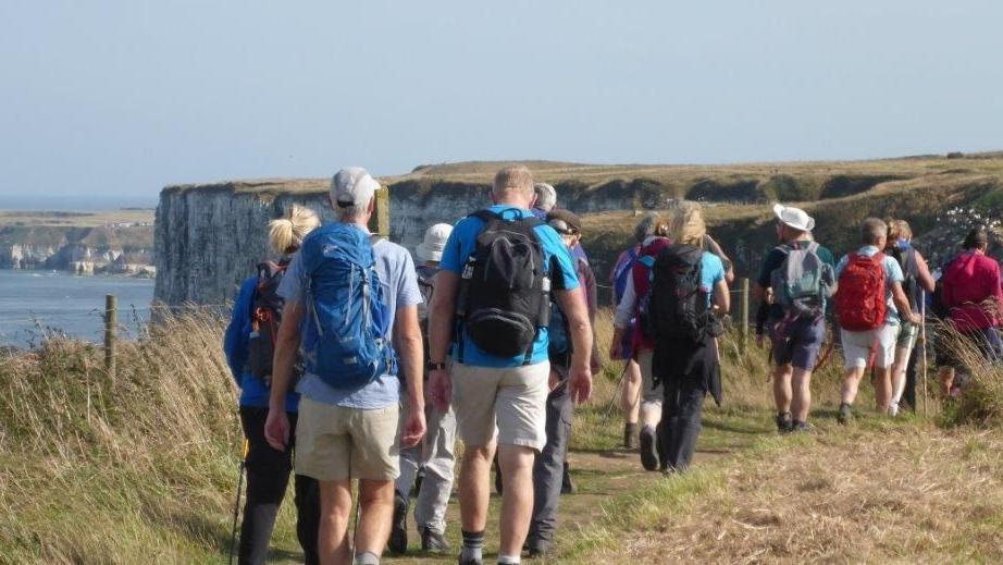 A group of men and women walk on a grass track towards the dramatic chalk cliffs of Flamborough Head. They are facing away from the camera and some are carrying rucksacks.