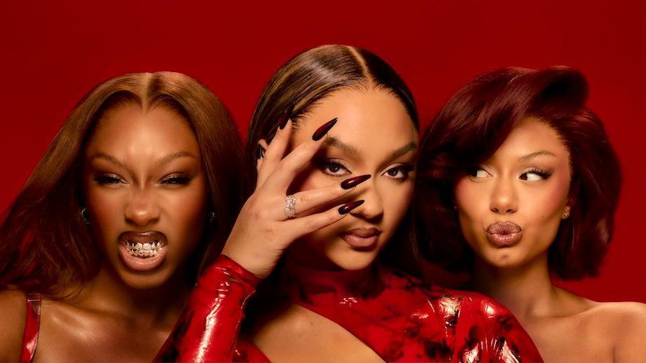 Promotional photo for the girl group Flo, showing all three members against a crimson red background. Singer Reneé Downer bares her teeth, bandmate Stella Quaresma poses with her hand partially obscuring her face, and Jorja Douglas pouts for the camera.