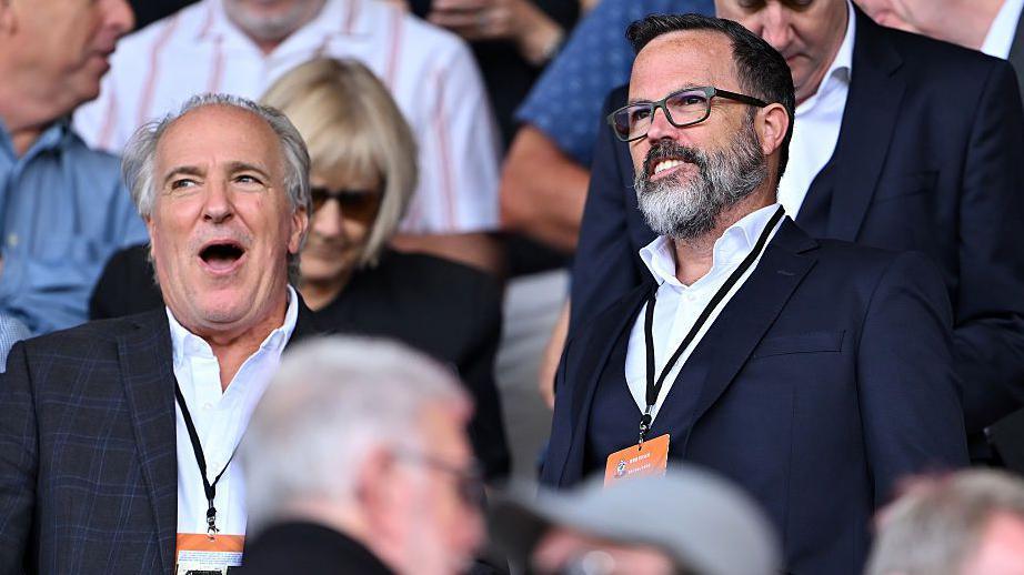Eric Allyn and Michael Williamson wearing dark suit jackets, open-necked white shirts and lanyards