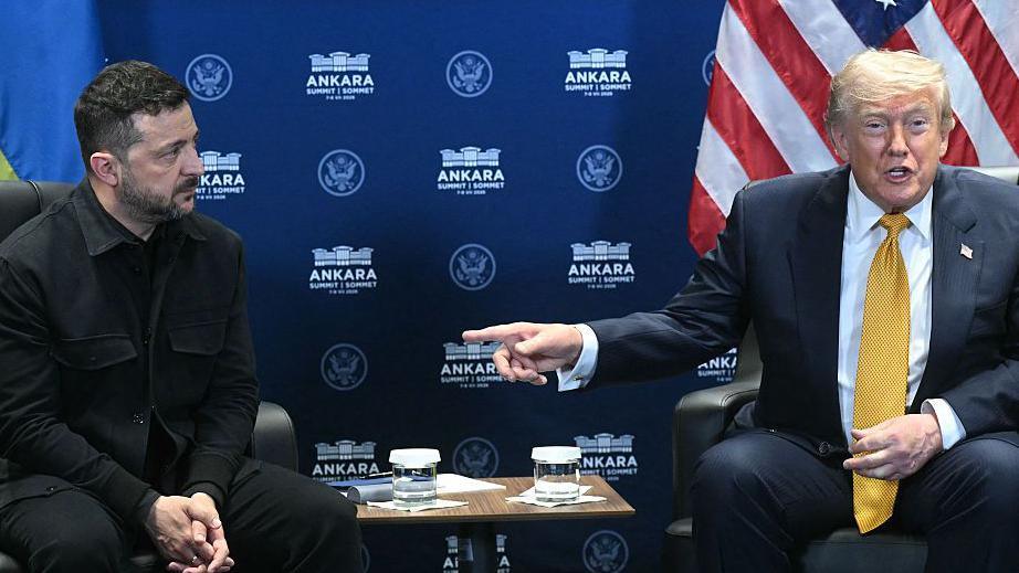 Zelensky and Trump seated at a news conference at the Nato summit in Ankara, Turkey. Trump is speaking to the cameras and pointing at Zelensky, who is looking towards him.
