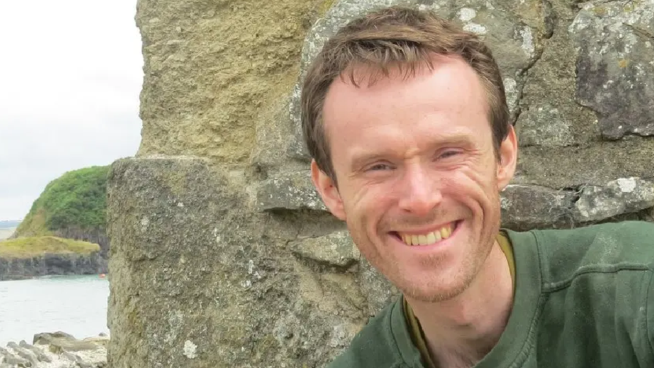 Carwyn Jones smiling looking at the camera. He is standing in front of a rock with the sea behind him. Carwyn is wearing a green sweatshirt and has short brown hair.