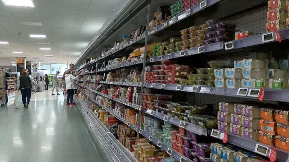 Food shelf inside a shop. The shelf is stocked with food items including dips. Shoppers are browsing the aisle.