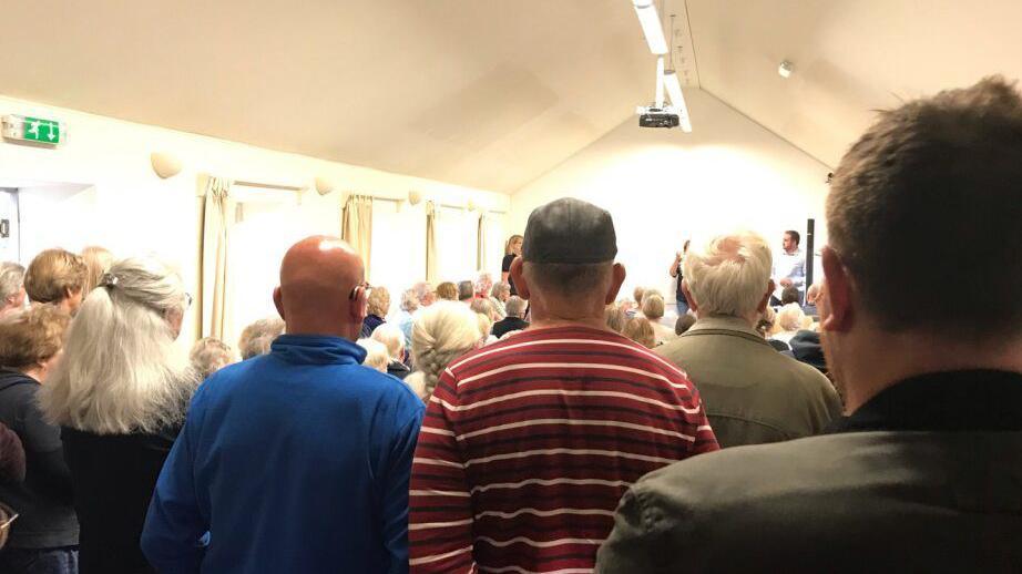 A packed out community hall with a number of people standing at the back.