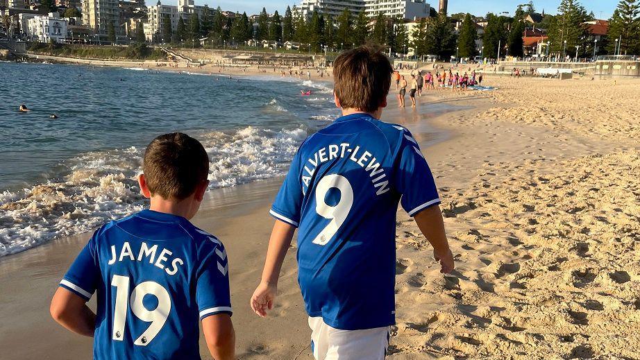 The backs of a young Mackenzie and his younger brother wearing blue Everton shirts with the names of players Dominic Calvert-Lewin and James Rodriguez while walking on a sandy beach in Australia.
