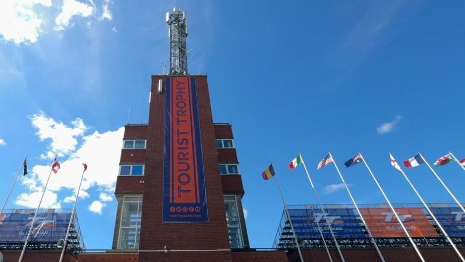 The tower at the grandstand, which is a red brick structure with an orange banner with Tourist Trophy written on it in blue lettering hanging down the front of it. Seating in the grandstand is either side of the tower and there is a row of flagpoles with international flag flying on them.