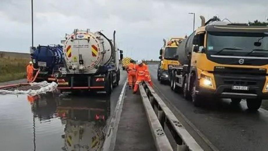 Two tankers on a road, with the left side flooded and water is being drained, while workers wearing orange overalls carry out work. There are also two other lorries on the right.