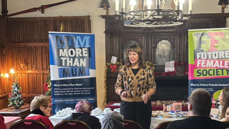 Ms Trotter is pictured standing in front of a seated crowd giving a talk. Behind her is a table with mugs and saucers as well as two information boards about the programme and Future Female Society.