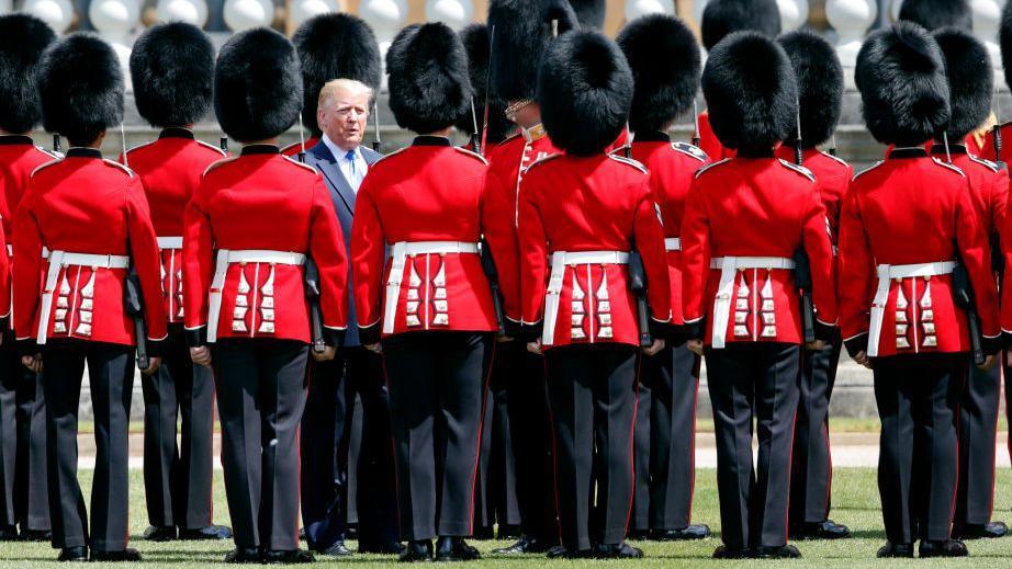 I veshur me një kostum blu dhe kravatë blu të zbehtë, Donald Trump inspekton një Gardë Nderi të formuar nga Rojet Grenadierë me veshje të kuqe në Pallatin Buckingham gjatë vizitës së tij shtetërore në Mbretërinë e Bashkuar në qershor 2019.