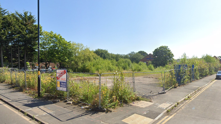 An area of land that is fenced off with metal fencing. There is a "sold" sign on the fence. Beyond the fence are overgrown plants, grass and weeds, as well as piles of gravel and dirt.