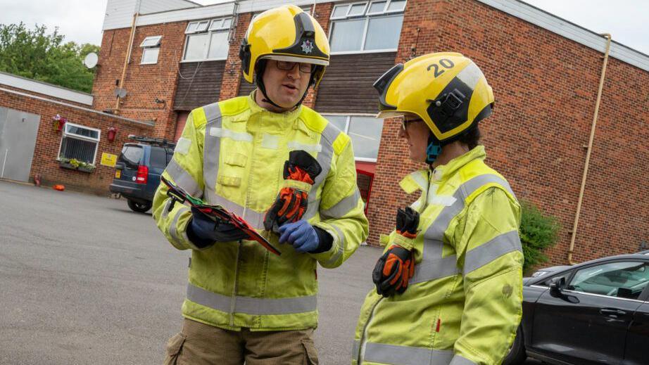 Two firefighters, in uniform, one of them is holding a ten-second 