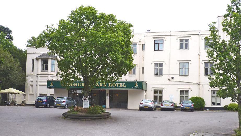 An exterior view of the three-storey white hotel, with the words Lyndhurst Park Hotel on a green sign above the entrance. Some cars are parked outside