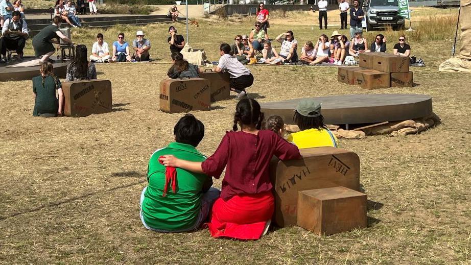 Children taking part in the outdoor performance, sitting with suitcases