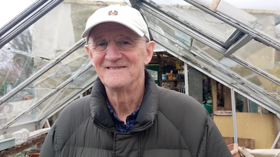 Tom Haire, with white cap and grey jacket, standing inside greenhouse with glass panels.