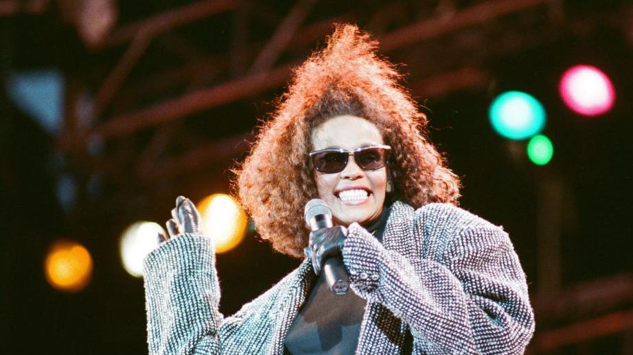 Whitney Houston singing at Freedomfest: Nelson Mandela 70th Birthday Celebration at Wembley Stadium in 1988. She is wearing an oversized grey cardigan, black leather gloves and dark glasses. 