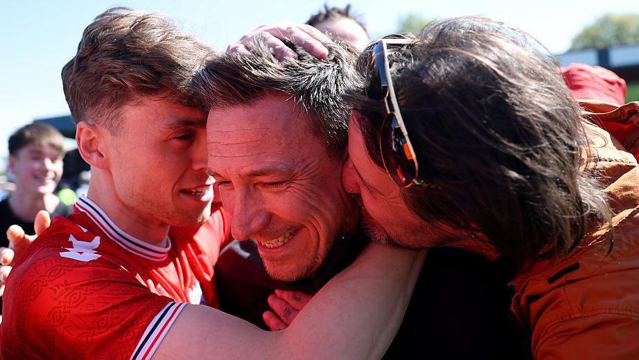 Stuart Maynard smiles as two York fans embrace him and put their foreheads next to Maynard's head while standing on the pitch