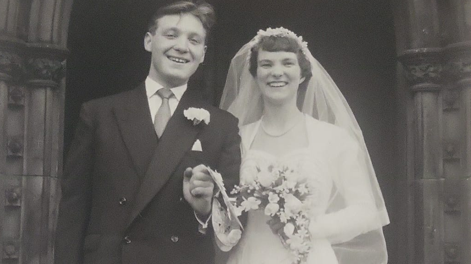 A young John and Barbara are standing together in a black and white image on their wedding day. Barbara is wearing a white wedding dress and holding flowers and John is dressed in a formal suit and holding her hand. They are both smiling at the camera.
