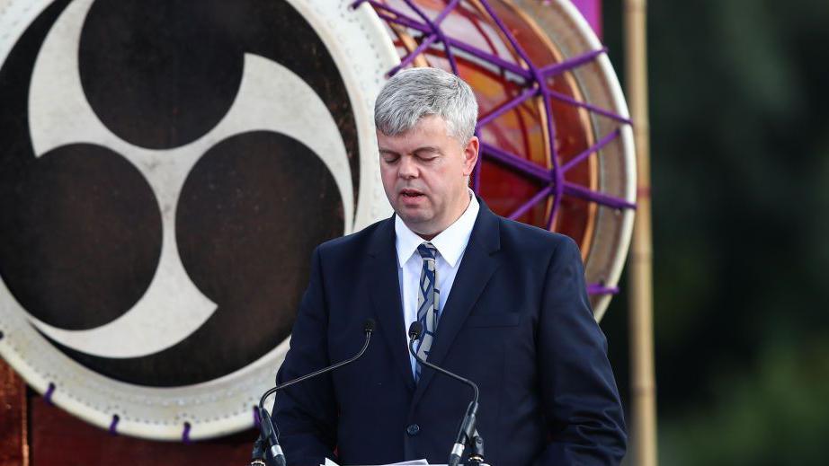 A man with grey hair and a blue suit and patterned blue tie stands in front of a microphone