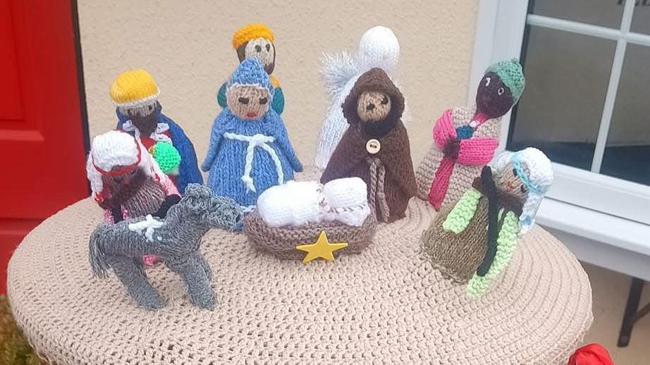 A collection of knitted figures includes Mary in a blue dress, Joseph in a brown cloak, an angel in white behind them, and baby Jesus in a crib in front of them. There is also a donkey along with other characters including the three wise men and a shepherd.