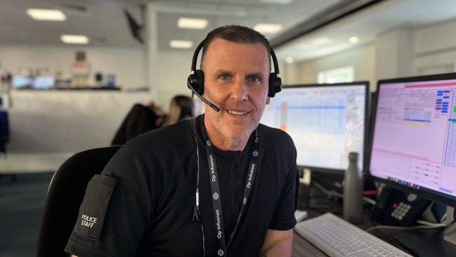 Jon Bridge, wearing a black police t-shirt and headset, sits at a desk in the York control room.