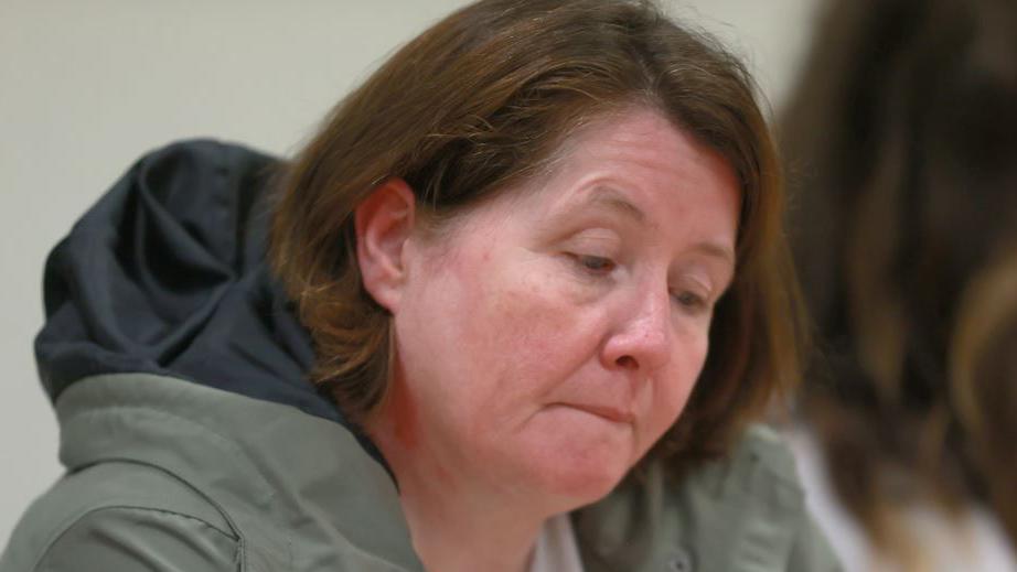 A woman with brown hair stares into space. She has brown hair and is wearing a green jacket