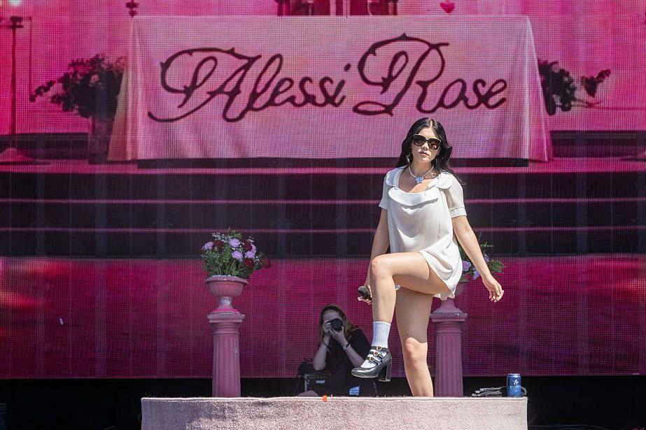 A woman with dark hair and sunglasses wearing a short white dress against a pink backdrop.