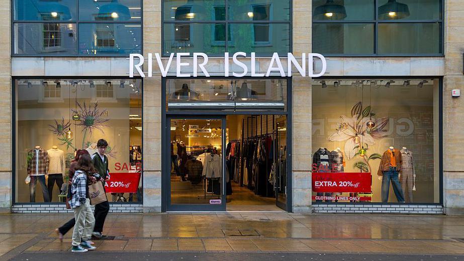 A group of young people walk past a River Island store with sale signs in the window
