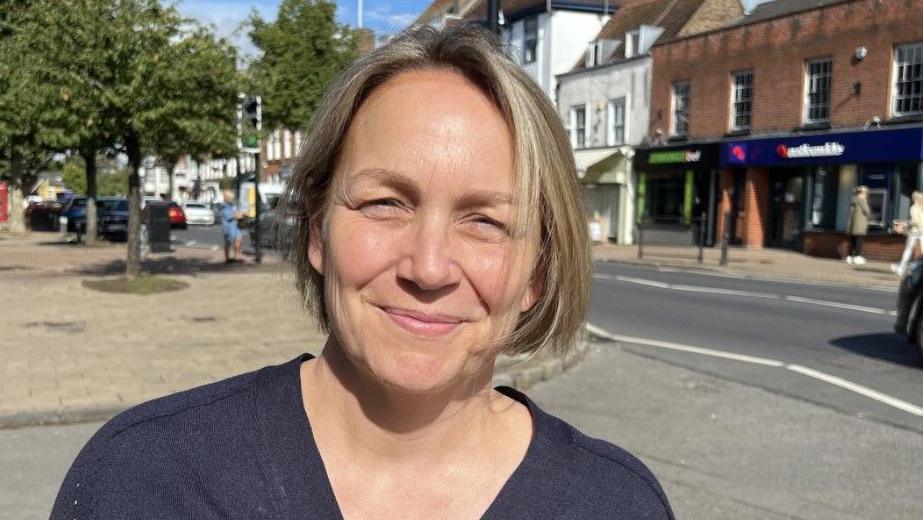 Kerry Gilroy is looking at the camera on a sunny day. She has a short blonde bob and is wearing a dark blue top. Shops can be seen in the background as well as people walking on a street and a car on the road
