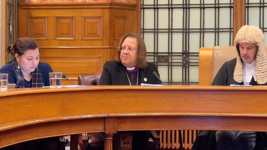 Bishop Tricia Hillas, is wearing a black suit jacket, with a purple shirt and white Church of England collar. She has brown shoulder length hair and wears glasses. She's sitting at the wooden legislative council bench, with the President of Tynwald in a traditional wig to the right, and Tanya August-Phillips MLC to the left.