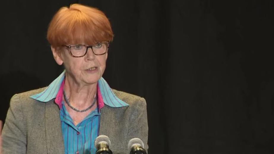 Dame Vera Baird with short ginger hair and wearing glasses, is photographed speaking at a press conference wearing a blue shirt and grey blazer