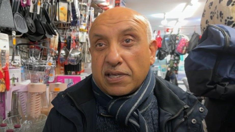 A man wearing a grey jumper as well as a navy blue coat and a striped navy blue scarf is stood in a shop. He is surrounded by items hung up such as cooking utensils and blankets for pets.