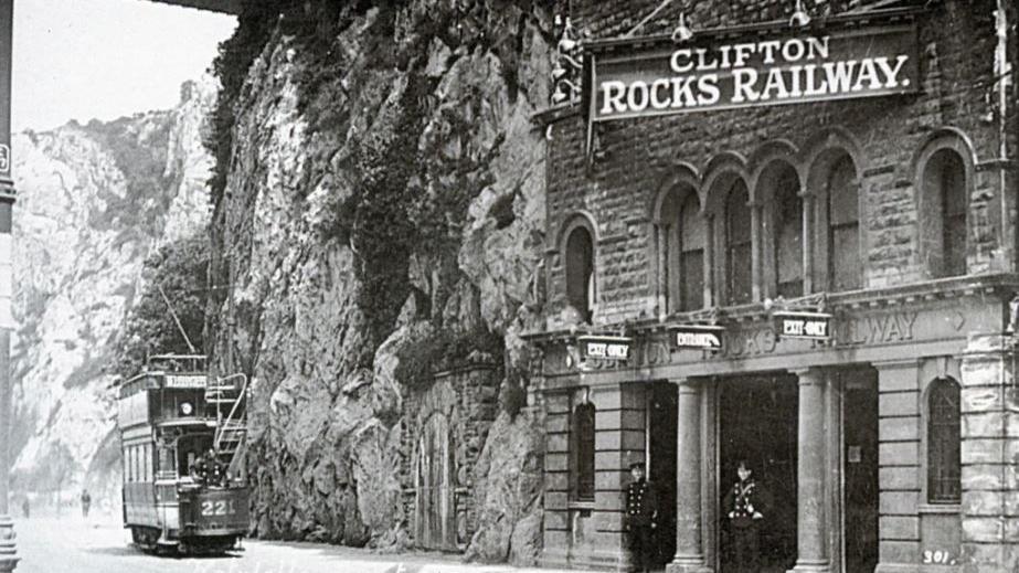 An archived black and white image of the Clifton Rocks Railway in operation. There is a large sign hanging above the tunnels and two members of staff are standing in the doorway. A tram car is driving past on what is now the Portway road.