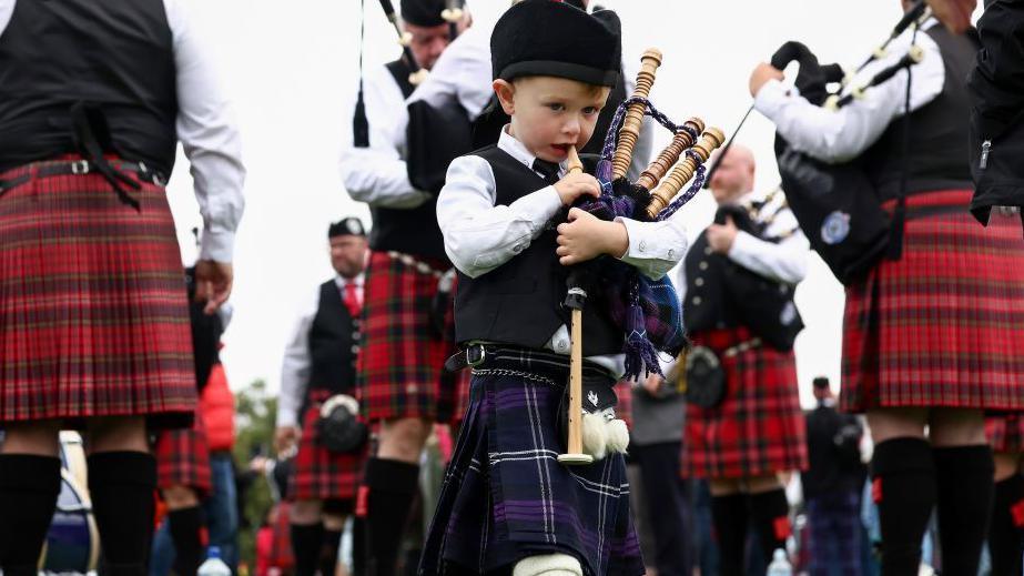 A young boy dressed as a piper blowing into mini bagpipes