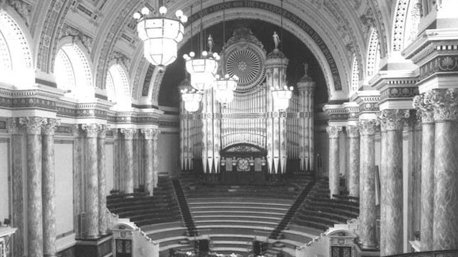 A large hall with a seating area and number of ornamental pillars against the wall. There are five chandeliers hanging from the ceiling and an organ at the back of the hall. 