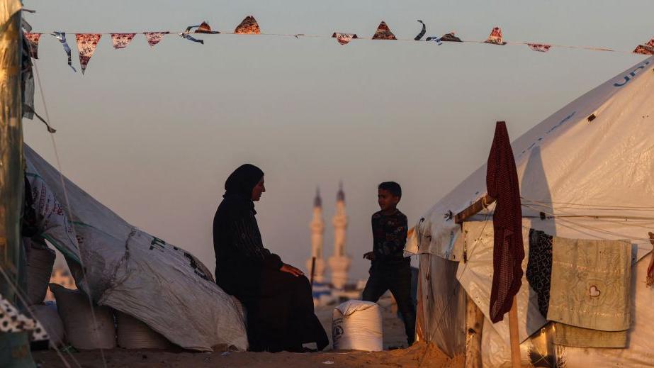 A boy walks past a Palestinian woman sitting at a camp for displaced people in Rafah, in the southern Gaza Strip on 11 March 2024