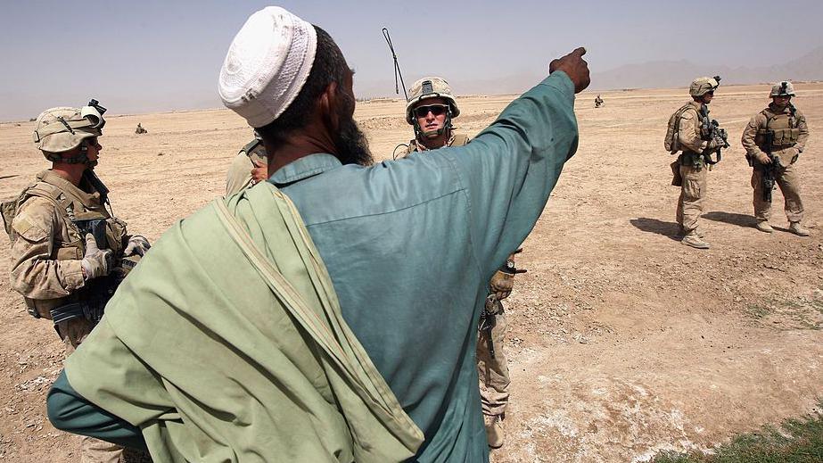 A man with his back to the camera dressed in traditional Afghan clothing speaks to a group of four US soldiers and an Afghan interpreter.