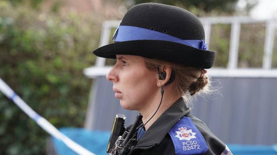 A close-up side profile of a female police officer in a blue and black uniform.  She is standing outside at the scene of Richard Langley's alleged murder.