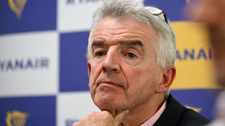 Michael O'Leary, who has short, grey hair and wears a black jacket over a pink shirt, sits at a table with his hand resting on his chin. A board with Ryanair logos are on a board behind him.