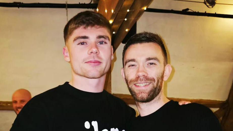 Leif Davis and Danny Campion stand together and smile at the camera. They both have short dark hair and wear dark T-shirts.