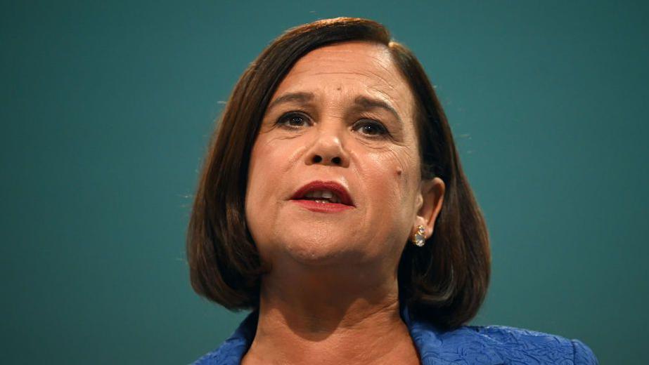 Close-up shot of Mary Lou McDonald. She has brown bobbed hair, red lipstick and earrings. The top of her jacket, which is blue, is visible. 