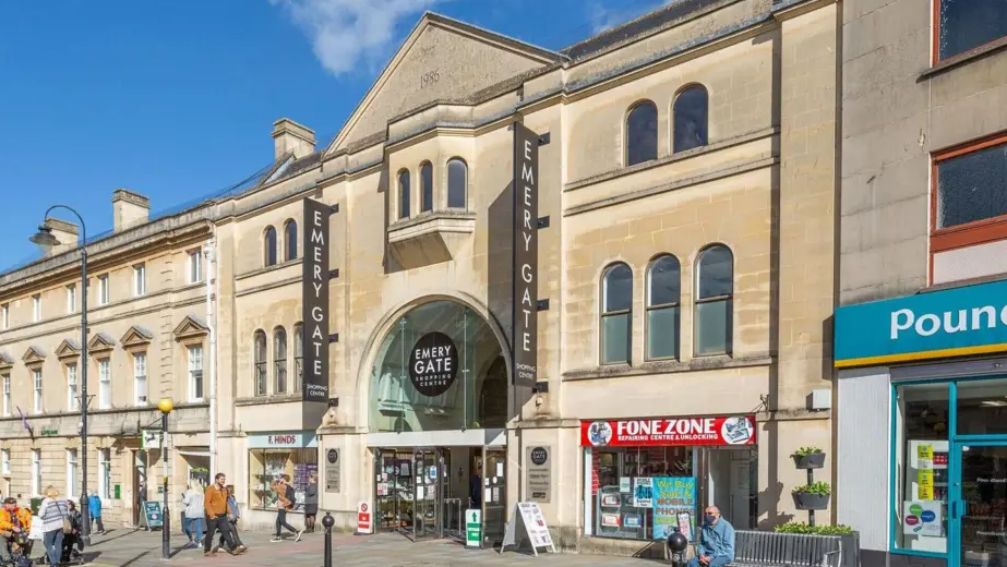 The exterior of Emery Gate shopping centre in Chippenham on a sunny day. It is a three story, stone building. Boards outside are advertising the shopping centre. The front of F. Hinds jewellery store and Lloyds Bank is visible on the left while Fone Zone and Poundland are on the right.