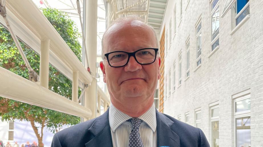 Matthew Hicks standing in a corridor next to an atrium, so it's very light. He is wearing a blue suit and blue patterned tie. He is smiling and wearing glasses. 