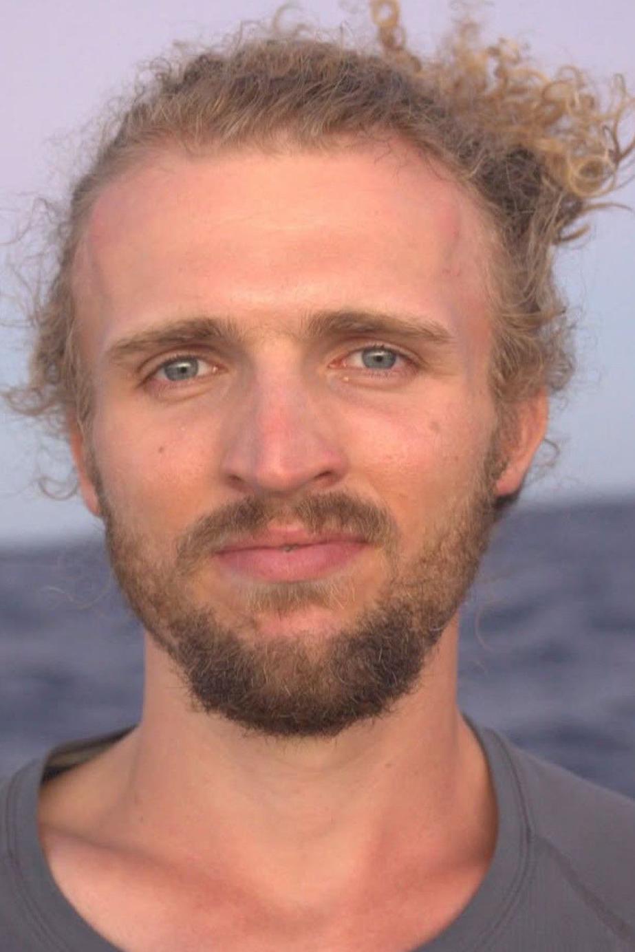 Lachlan has blonde wavy hair and a short beard. He is wearing a grey t-shirt and has the sea behind him.