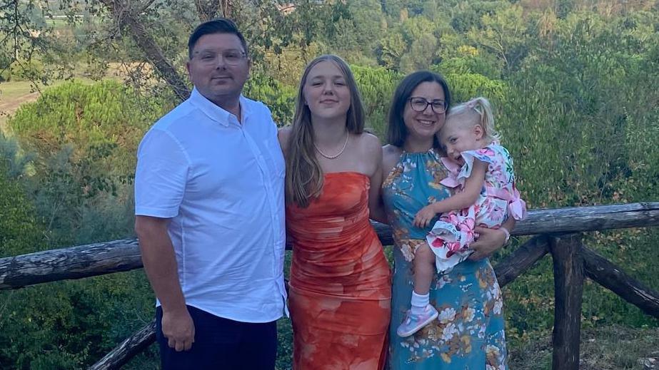 A family of four - a mother and father with their teenager daughter and four-year-old daughter - stand arm in arm smiling at the camera. The woman and her daughters are wearing summer dresses while the husband wears a white shirt with black shorts. Behind them is a wooden fence and green foliage.