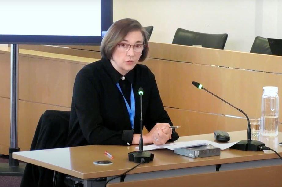 Eleanor Grey KC, wearing a black blouse with a blue ribbon around her neck, is sitting at a desk with two microphones in front of her.  