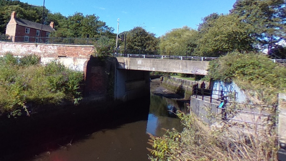 Photo of a low concrete bridge over small river on clear blue day. There is a large house in the background and the area is surrounded by trees and bushes. 