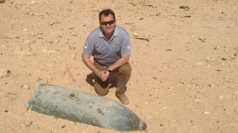 Mr Remfrey is crouched down next to a mine in an arid environment. He is wearing sandy coloured shoes and trousers and a grey polo shirt and sun glasses. He has black hair.